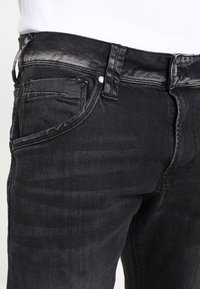 Pantalones vaqueros de mezclilla negra con un acabado ligeramente desgastado, que presentan bolsillos delanteros, cremallera y detalles de herrajes metálicos. Diseño clásico de cinco bolsillos.