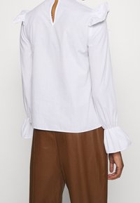 Blusa bianca in cotone con accentuazioni di spalle a volant, maniche lunghe con polsini arricciati e dettaglio a occhio di bue sul retro.