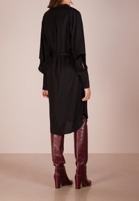 Vestido negro de manga larga con cintura ceñida, fabricado en un material suave, cuello alto y aberturas laterales, combinado con botas de cuero borgoña hasta la rodilla.