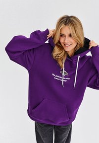 Fialová mikina s kapucí a přední kapsou, bílé šňůrky a potisk s textem. Měkká textura, oversized střih a vroubkované manžety.