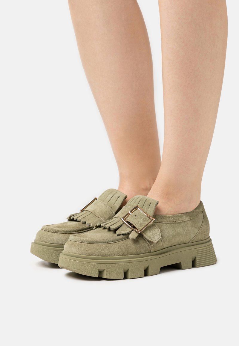 Vêtements pour les jambes et mocassins en daim vert olive avec semelles épaisses à rainures, languettes à franges et grandes boucles carrées dorées sur fond blanc.