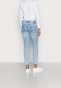 Světle modré džíny z denimu s úzkým střihem, které mají dvě zadní kapsy a béžovou koženou záplatu na pásku, doplněné bílými teniskami.