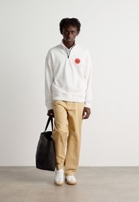 White zippered sweatshirt with red logo, beige trousers, and white sneakers. Model holds a black tote bag. Simple and modern design.