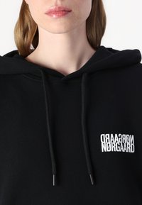 Hoodie noir avec cordons de serrage réglables, arborant un logo imprimé blanc sur le côté gauche. Fabriqué à partir d'un tissu doux et texturé.