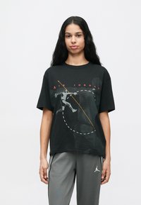 Νεαρή γυναίκα με μακριά μαύρα μαλλιά, φορώντας μαύρο μπλουζάκι Air Jordan με γραφικά και γκρι παντελόνι Air Jordan, στέκεται μπροστά σε λευκό φόντο.