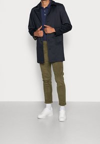 Mörkblå trenchcoat över en mörkblå polo, tillsammans med olivgröna byxor och vita sneakers. Tyget är slätt och har en klassisk knapdesign.