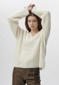 Weißer strukturierter Strickpullover mit V-Ausschnitt und lockeren Ärmeln, kombiniert mit braunen Lederhosen. Weicher Stoff und lässige Passform.