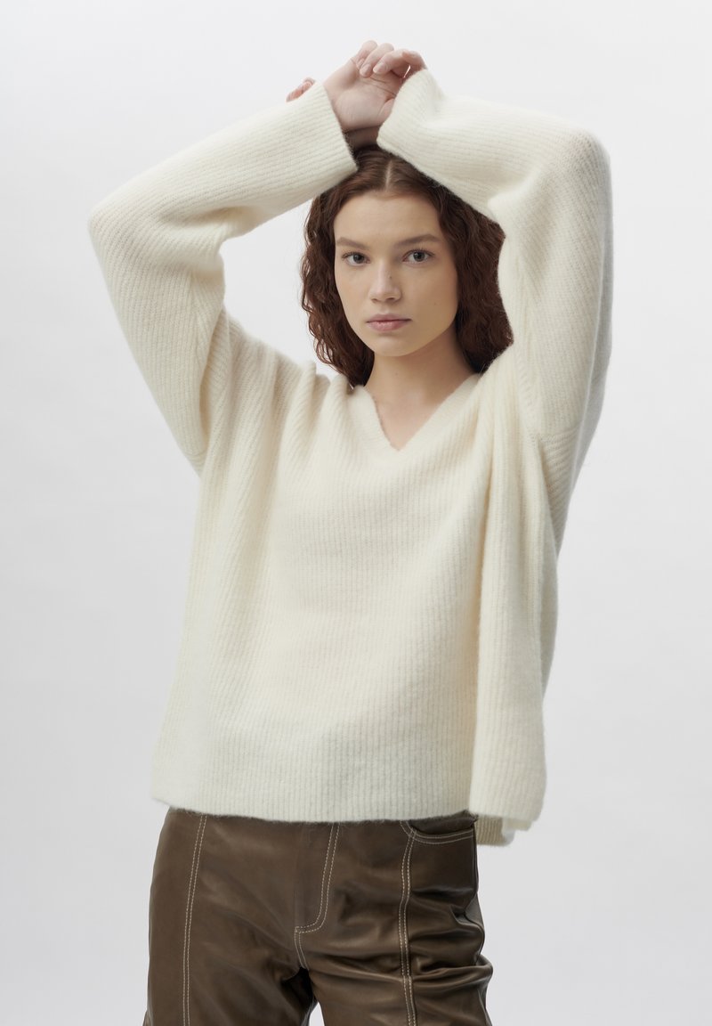 Weißer strukturierter Strickpullover mit V-Ausschnitt und lockeren Ärmeln, kombiniert mit braunen Lederhosen. Weicher Stoff und lässige Passform.