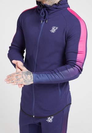 Mężczyzna w dresie w gradient od granatu do różu z kapturem, stojący z złączonymi rękami, pokazujący tatuaże na palcach i nadgarstku.