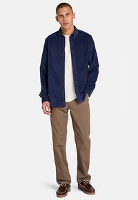 Navy button-up shirt over a cream T-shirt, paired with light brown pants and dark brown boat shoes. Casual design, cotton material.