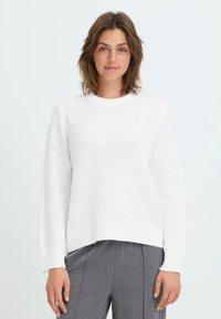 White knitted sweater with a textured pattern, ribbed neckline and cuffs, and a cropped design. Paired with grey trousers.