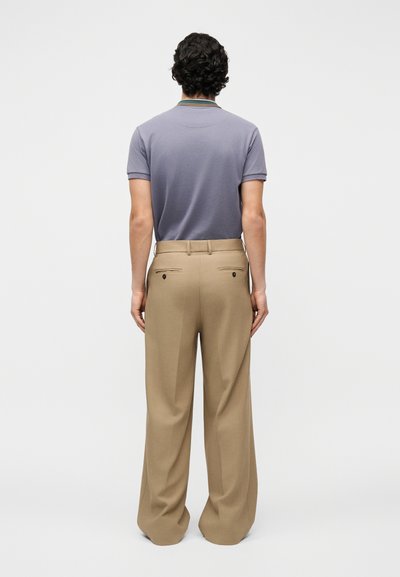 Homme aux cheveux bouclés et foncés portant un polo gris à manches courtes rentré dans un pantalon beige à jambes larges, debout de dos.