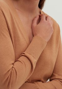 Jasnobrązowy dziergany kardigan z dekoltem w kształcie litery V. Posiada długie rękawy i miękką fakturę, z detalem w postaci guzika przy talii.