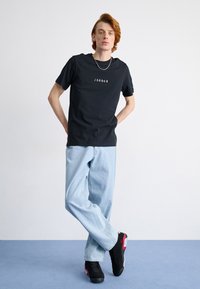 Juodas trumpas rankovėmis marškinėliai su "JORDAN" logotipu, derinami su šviesiai mėlynais laisvais džinsais ir juoda avalyne su raudonais akcentais.