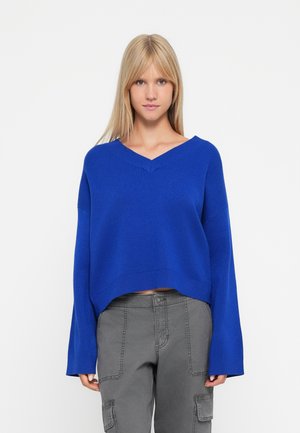 Maglione blu cobalto a maniche lunghe con scollatura a V e orlo a coste. Indossato con pantaloni cargo grigi. Tessuto morbido.