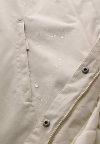 Beige waterdichte jassenstof met zichtbare waterdruppels, voorzien van een ritsluiting en drukknoopdetails langs de rand.