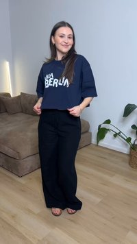 Νεαρή γυναίκα χαμογελαστή, φορώντας μια navy oversized cropped μπλούζα με το κείμενο "LALA BERLIN" και μαύρο παντελόνι με φαρδιά μπατζάκια, στέκεται σε ένα μοντέρνο σαλόνι.