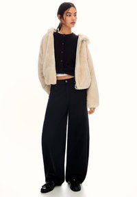 Veste en fausse fourrure beige avec capuche, haut noir à boutons, pantalon noir à jambes larges et chaussures noires brillantes. Lignes épurées et style superposé.