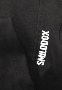 Černá mikina vyrobená z měkké tkaniny, která obsahuje velké bílé logo "SMILODOX" na boku a jemnou strukturu.