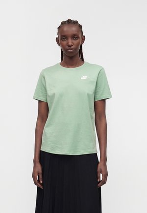 Fonott hajjal rendelkező nő, aki világoszöld Nike pólót és fekete pliszírozott szoknyát visel, egyszínű világos háttér előtt áll.