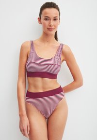 Vrouw die tegen een witte muur staat, gekleed in een maroon en wit gestreepte sportbeha en bijpassende high-waisted onderbroek, met ontspannen armen.