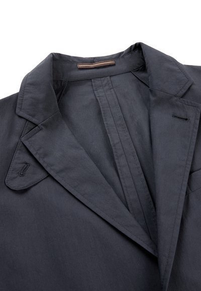 Donkergrijze blazer met een reverskraag met inkeping, knoopsgatdetails en een kleine stoffen lus aan de binnenkant van de achterkant van de hals, tegen een witte achtergrond.
