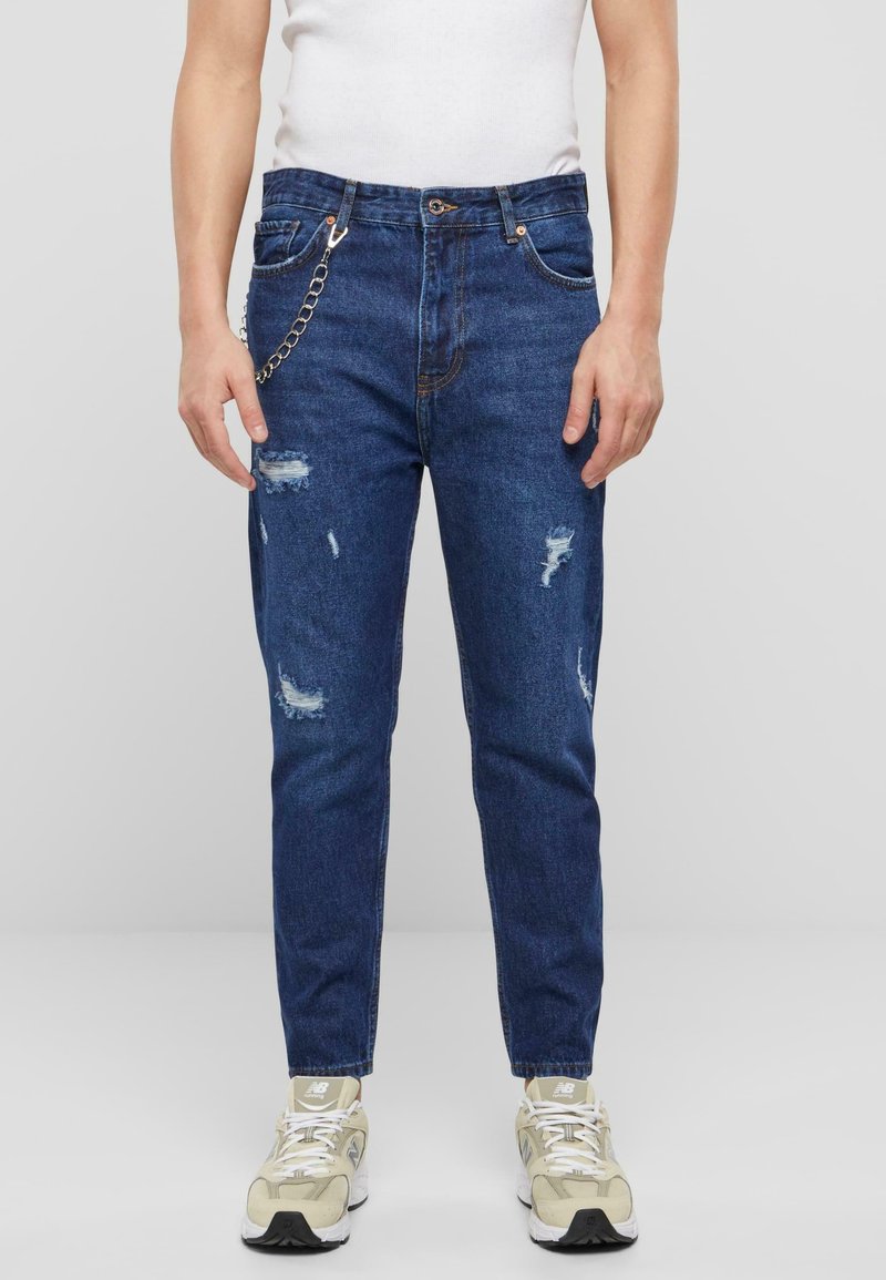 Personne portant un jean bleu délavé avec un détail chaîne, une chemise blanche et des baskets beiges, debout devant un fond uni gris clair.