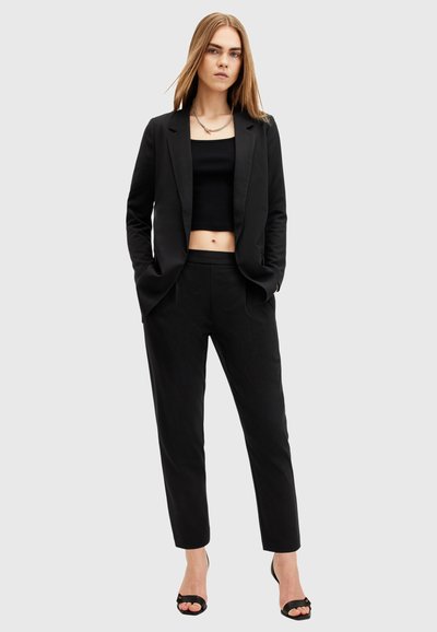 Blazer noir et top court associés à un pantalon noir ajusté. Tissu lisse avec un design cintré, complété par des talons ouverts.