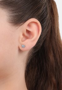 Kleine herzförmige Ohrringe mit hellblauen glitzernden Steinen, die in einer Metallbasis gefasst sind, getragen am Ohrläppchen einer Frau.