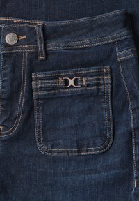 Poche de jean en denim bleu foncé avec un bouton en métal et des coutures cousues, avec un petit logo décoratif en métal sur le rabat de la poche.