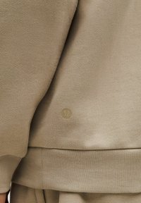 Beige collegepaita, jossa on ribbaketju helmoissa ja hihansuissa, ja jossa on pieni, pyöreä logo hillityssä kultaisessa sävyssä. Pehmeä, teksturoitu kangas.