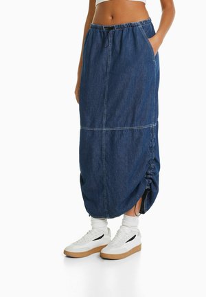 Kvinde iført en mørkeblå denim lang nederdel med detaljeret sidesamling, hvide sneakers og hvide sokker, hænderne i lommerne.