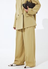 Bēša pārmērīga mētelis ar dubultu pogu dizainu, apvienots ar atbilstošām platām biksēm un norādītiem apaviem ar govs rakstu.