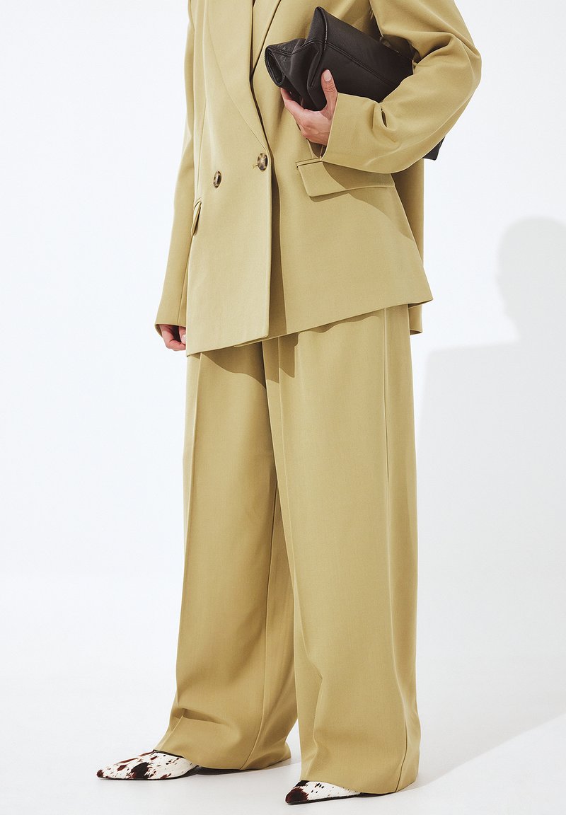 Bēša pārmērīga mētelis ar dubultu pogu dizainu, apvienots ar atbilstošām platām biksēm un norādītiem apaviem ar govs rakstu.