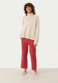 Crème turtleneck trui met geribbelde zoom; rode corduroy broek; zilveren ballerina's. Krullend haar omlijst het gezicht. Eenvoudige, relaxte pasvorm.