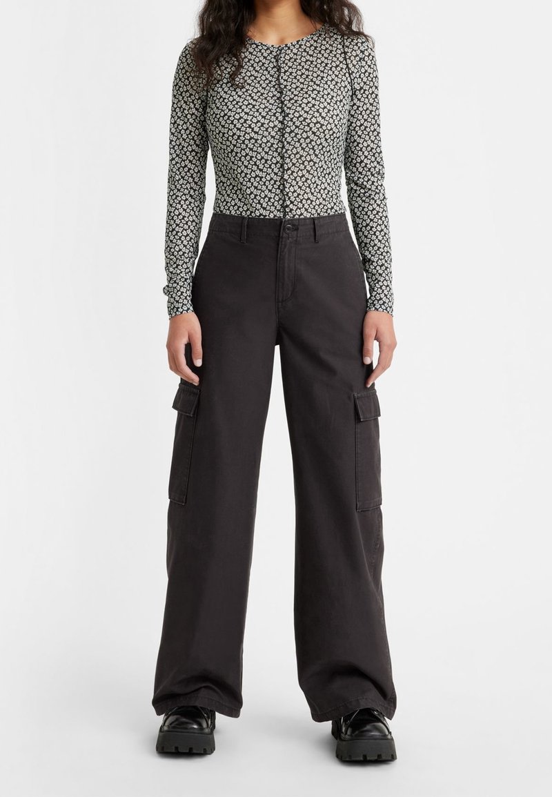 Femme portant un haut à manches longues noir et blanc à motif floral, un pantalon cargo noir à jambes larges et des chaussures noires épaisses sur un fond uni.