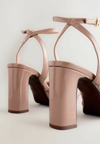 Nude högklackade sandaler med glänsande yta, korsremdesign och kraftig blockklack, som visar en slät textur och subtila guldfärgad detaljer.