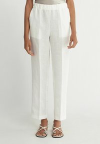 Weiße Leinenhose mit einem elastischen Bund, zwei Seitentaschen und geradem Beinverlauf. Dazu werden schlichte Sandalen mit Riemen getragen.