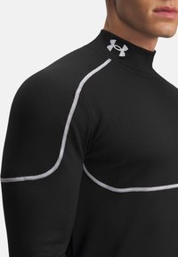Sort atletisk langærmet skjorte har hvide sting langs skuldrene, en høj krave og et Under Armour-logo ved halsudskæringen.