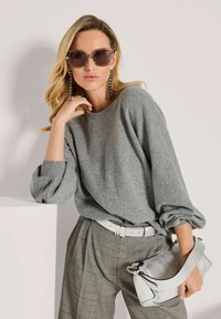 Eine Frau trägt übergroße Sonnenbrillen, einen grauen Pullover mit Puffärmel, karierte Hosen, hält eine silberne Handtasche und trägt lange hängende Ohrringe.