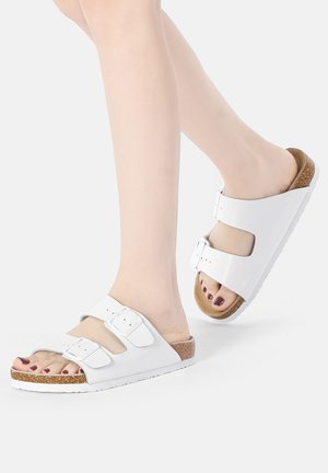 White slide sandals with two adjustable straps, cork footbed, rubber outsole, and a smooth finish. Designed for comfort and durability.
