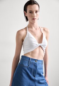 Weißes, geknotetes Crop-Top mit Halsbandträgern, kombiniert mit einem hoch taillierten blauen Denim-Rock. Auffällige Nähte und goldfarbene Hardware-Beschläge sind sichtbar.