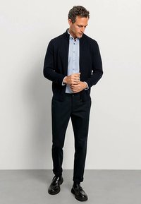 Navy knit cardigan over a light blue button-up shirt, paired with dark trousers and black shoes. Smooth texture and casual design.