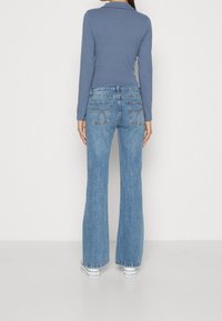 Kvinna som står vänd bort, iklädd en blå långärmad tröja, högmidjade ljusblå utsvängda jeans och vita platåskor mot en enkel bakgrund.