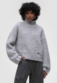 Šedý oversized svetr s vysokým límcem. Měkká, texturovaná tkanina a volné rukávy. Obsahuje detail loga na lemu.