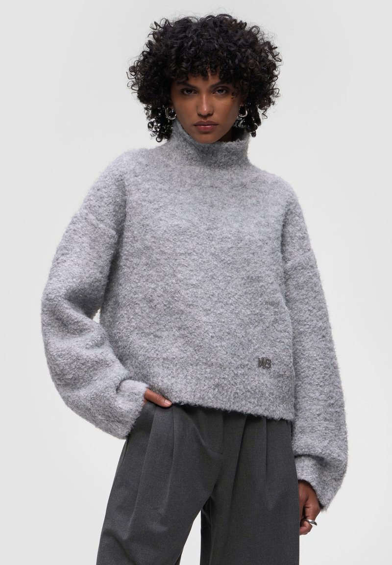 Šedý oversized svetr s vysokým límcem. Měkká, texturovaná tkanina a volné rukávy. Obsahuje detail loga na lemu.