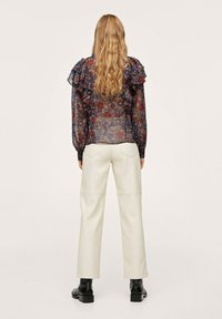 Femme aux longs cheveux ondulés portant un chemisier floral transparent avec des volants, un pantalon droit blanc cassé et des bottines noires, de dos.