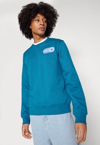 Türkiser Sweatshirt mit langen Ärmeln und rundem Hals. Verfügt über einen weißen Kragen, ein blauer Logo-Patch auf der Brust und gerippte Bündchen.