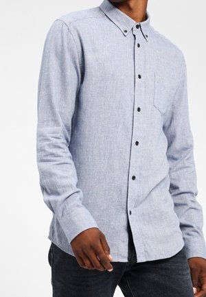 Hombre con una camisa de manga larga azul claro, abotonada, con un bolsillo en el pecho y jeans oscuros, visto desde la mitad del torso hasta el cuello.