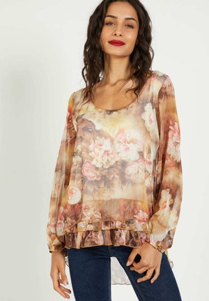 Mujer con cabello rizado y oscuro, que lleva una blusa transparente de manga larga con estampado floral y jeans azul oscuro, de pie frente a un fondo liso.